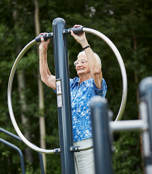 An elderly woman uses flexibility equipment at an outdoor fitness gym area.